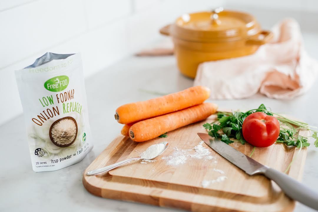 Low-FODMAP-Zwiebelersatz neben einem Schneidebrett mit Karotten, Tomate, Kräutern und Kochutensilien.