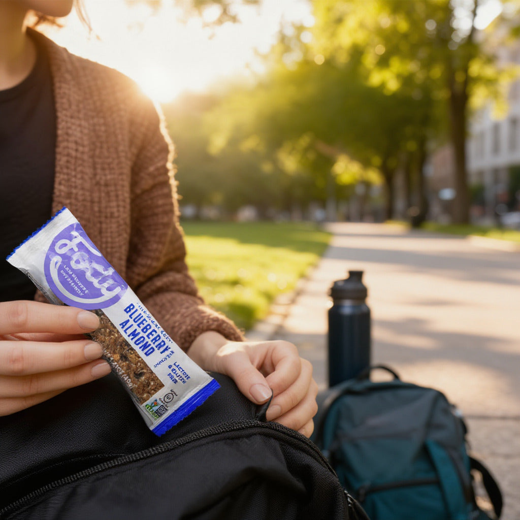 Frau im Park, mit Riegel in der Hand, neben ihr ein Rucksack und eine Trinkfalsche.