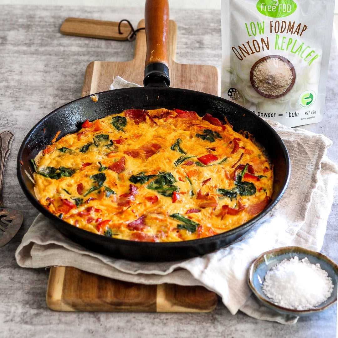 Gemüsefrittata in schwarzer Pfanne auf einem Holzbrett, daneben ein Beutel mit Low-FODMAP-Zwiebelersatz.