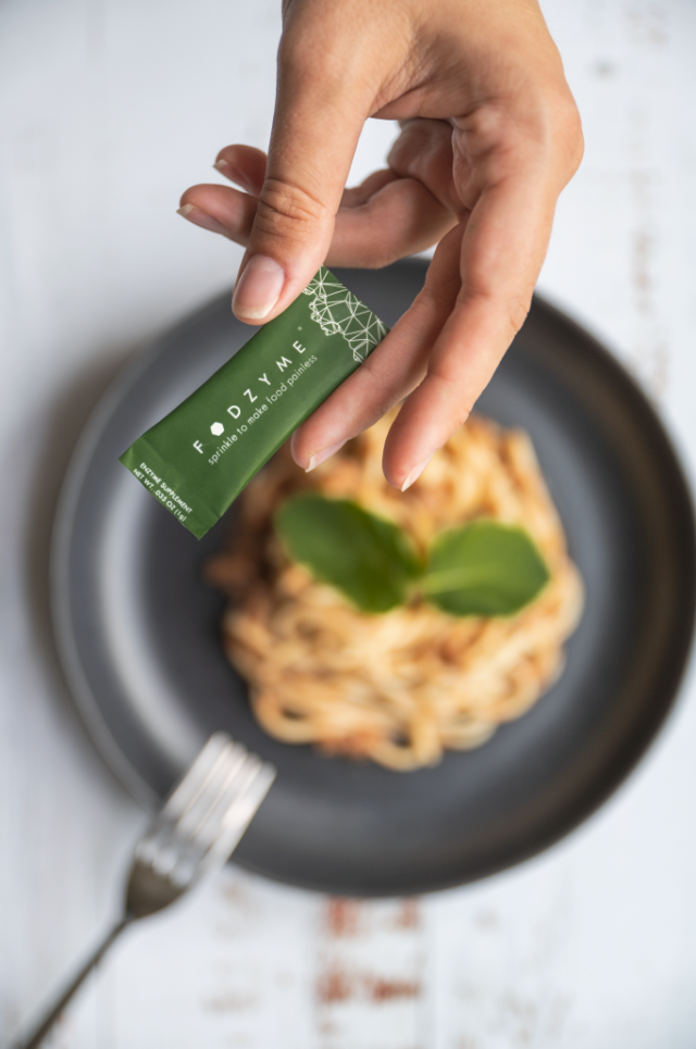 Hand hält ein Fodzyme -Päckchen über einem Teller Pasta.