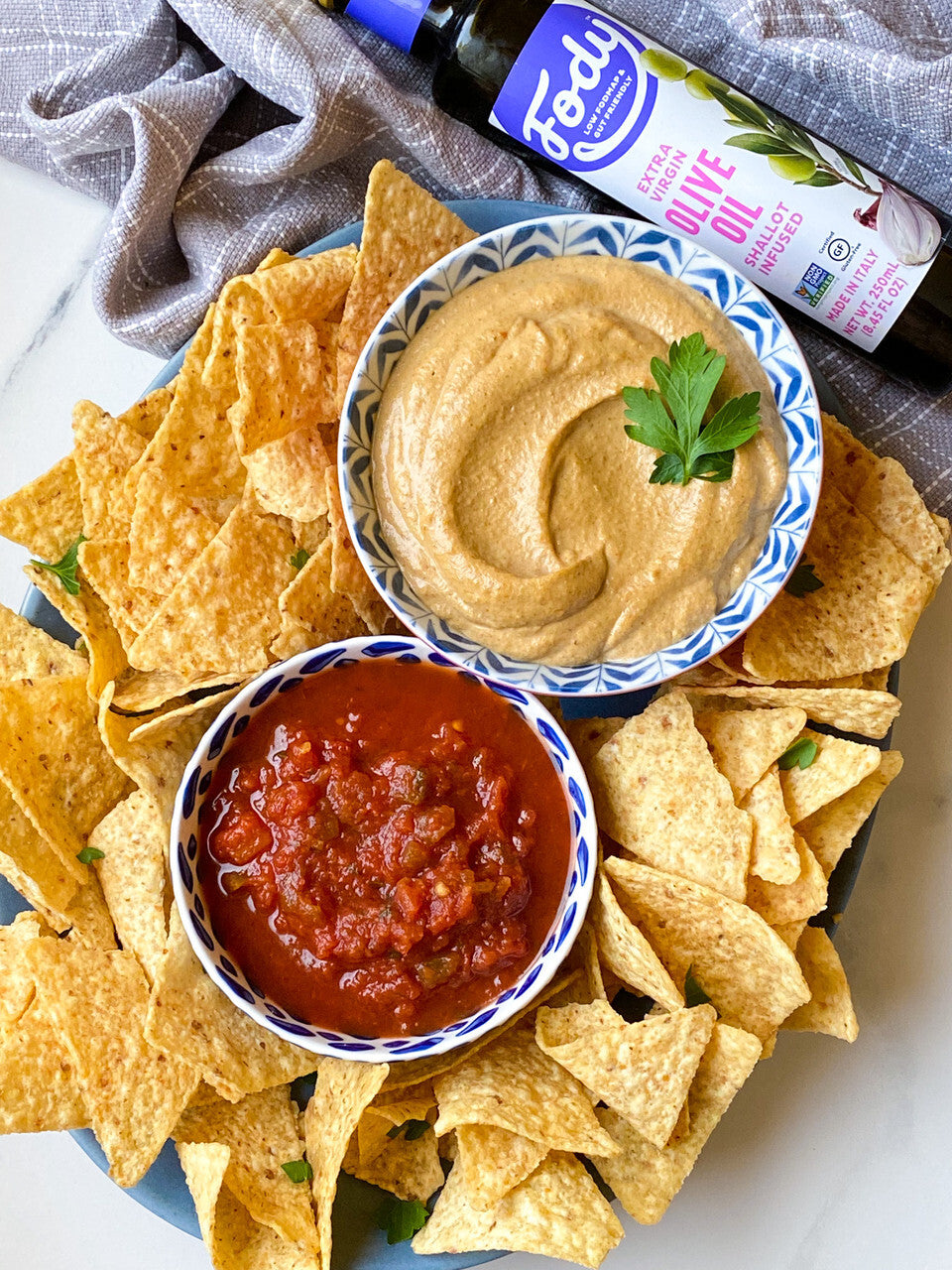 Tortilla-Chips auf einem Teller mit Hummus, Tomatensalsa und einer Flasche Fody-Olivenöl.